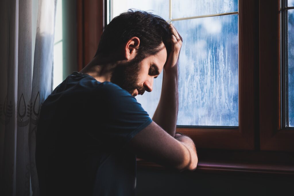 man struggling with heroin addiction looking out of a window with head down